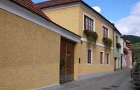 Gelbes Haus mit roten Dachziegeln und Blumenkästen an den Fenstern in einer schmalen Straße.