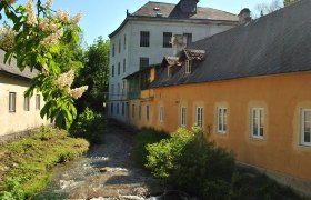 Historisches Geb&auml;ude mit Bach und bl&uuml;hendem Baum im Vordergrund.