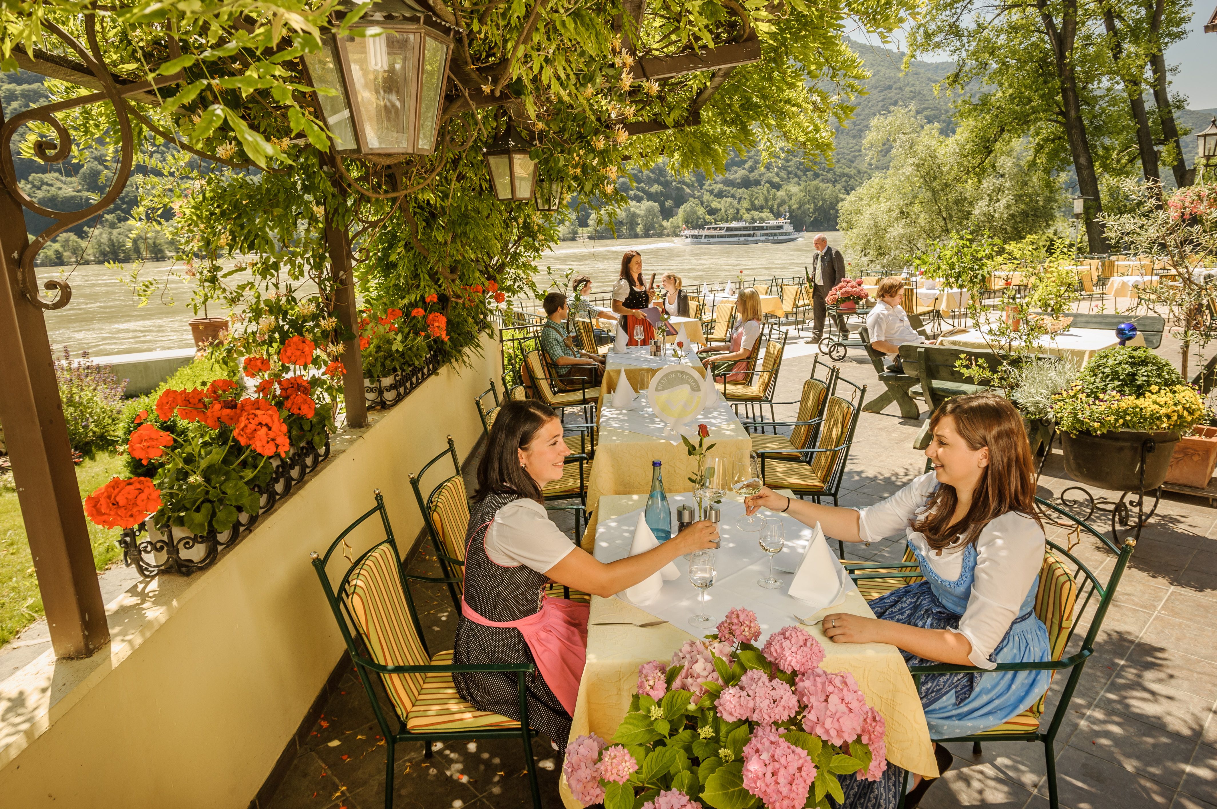 Menschen sitzen in einem Gartenrestaurant mit Blick auf einen Fluss, umgeben von Blumen und Bäumen.