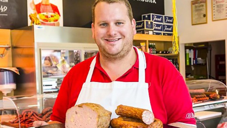 Ein Metzger in rotem Hemd und wei&szlig;er Sch&uuml;rze h&auml;lt verschiedene Wurstwaren in einer Fleischerei.