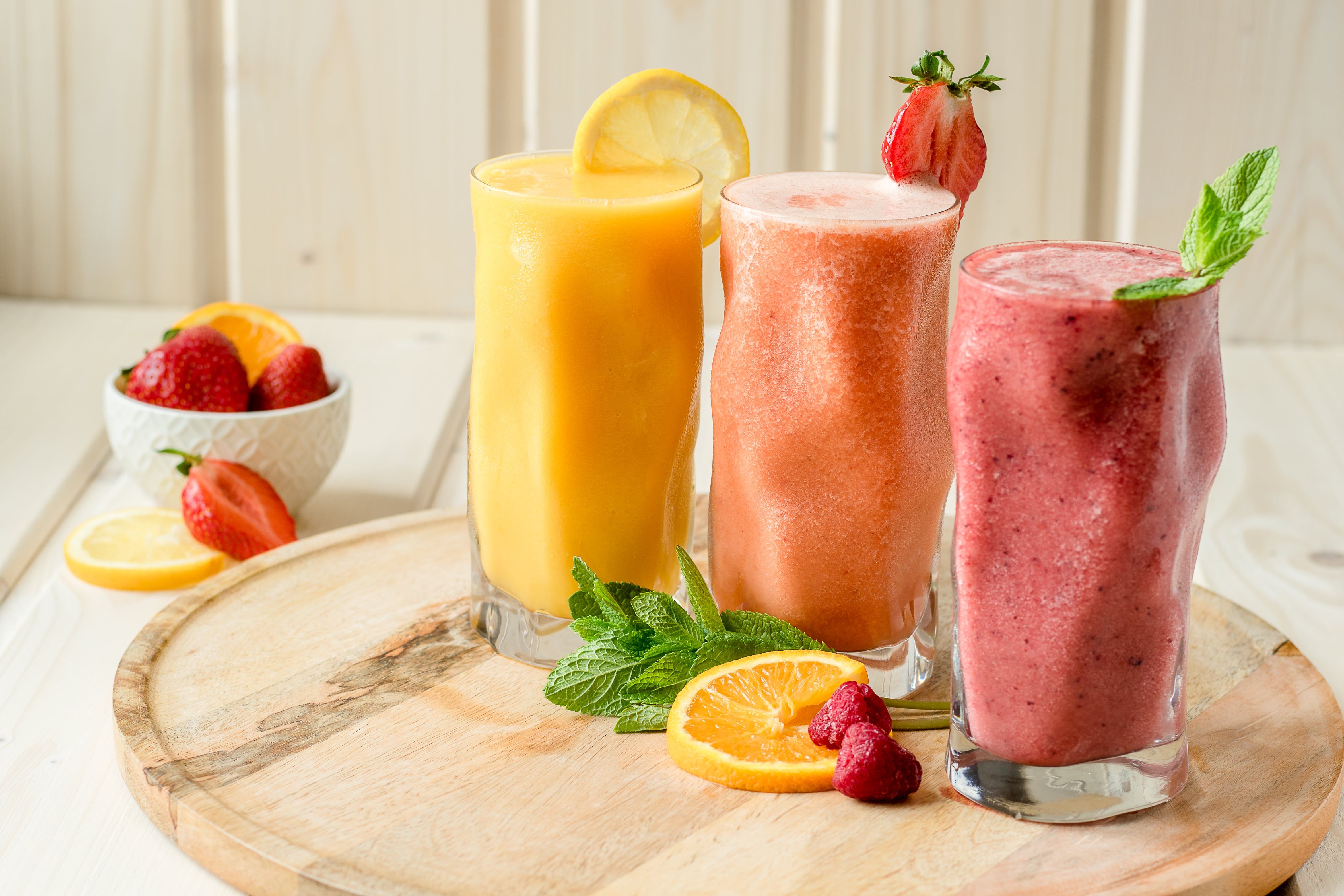 Drei Smoothies in Gläsern auf einem Holzbrett, dekoriert mit Obst und Minze.