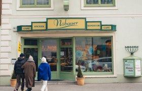 In der winterlichen Atmosphäre schlendern Besucher entspannt zur charmanten Konditorei, die mit ihren einladenden Fenstern und dem warmen Licht einladend wirkt. Der Duft frisch gebackener Leckereien zieht durch die kalte Luft und verspricht süße Genüsse, während die schneebedeckten Berge im Hintergrund eine malerische Kulisse bieten.