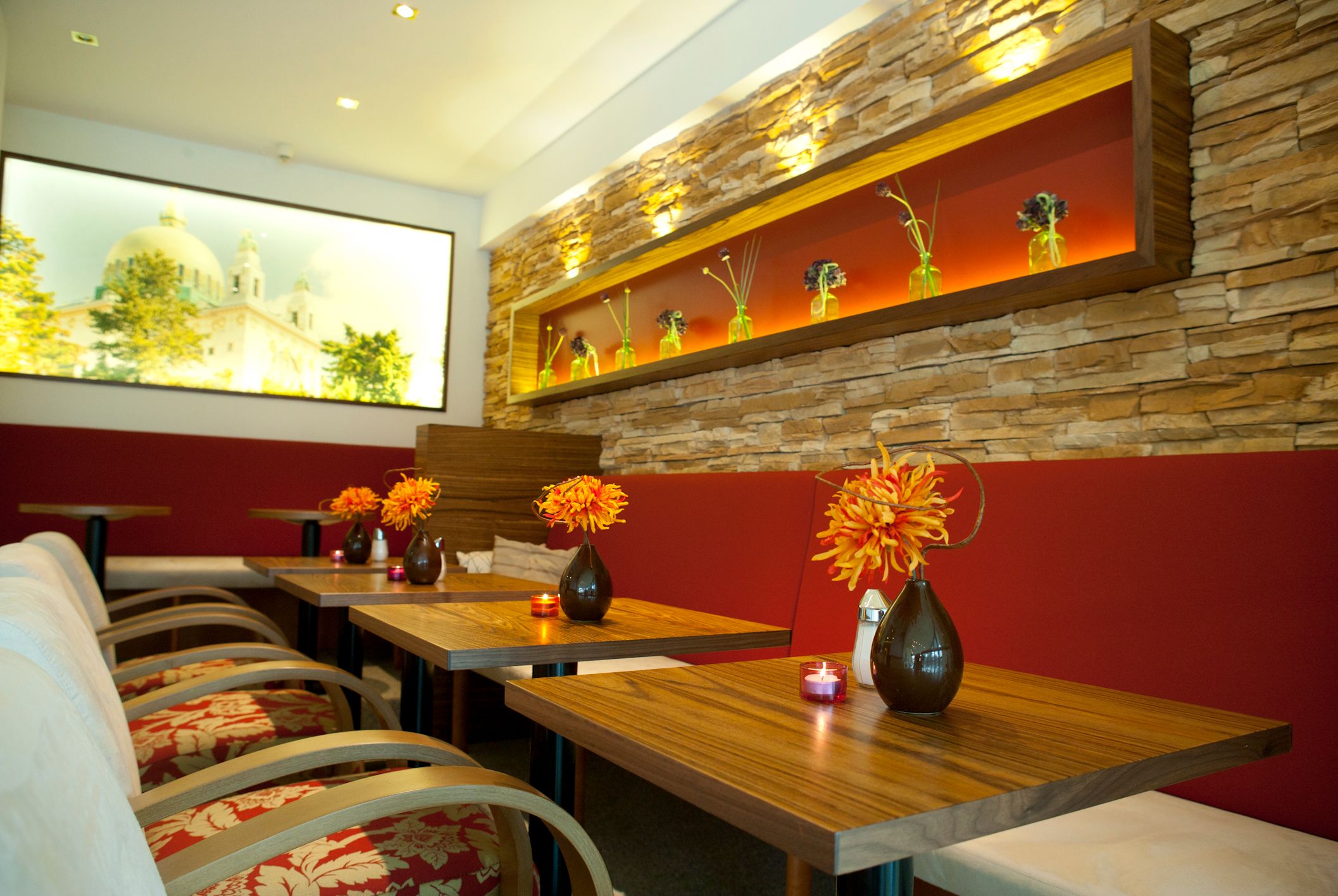 Innenansicht einer stilvollen Tagesbar mit Holztischen, roten Sitzbänken und dekorativen Blumenvasen.