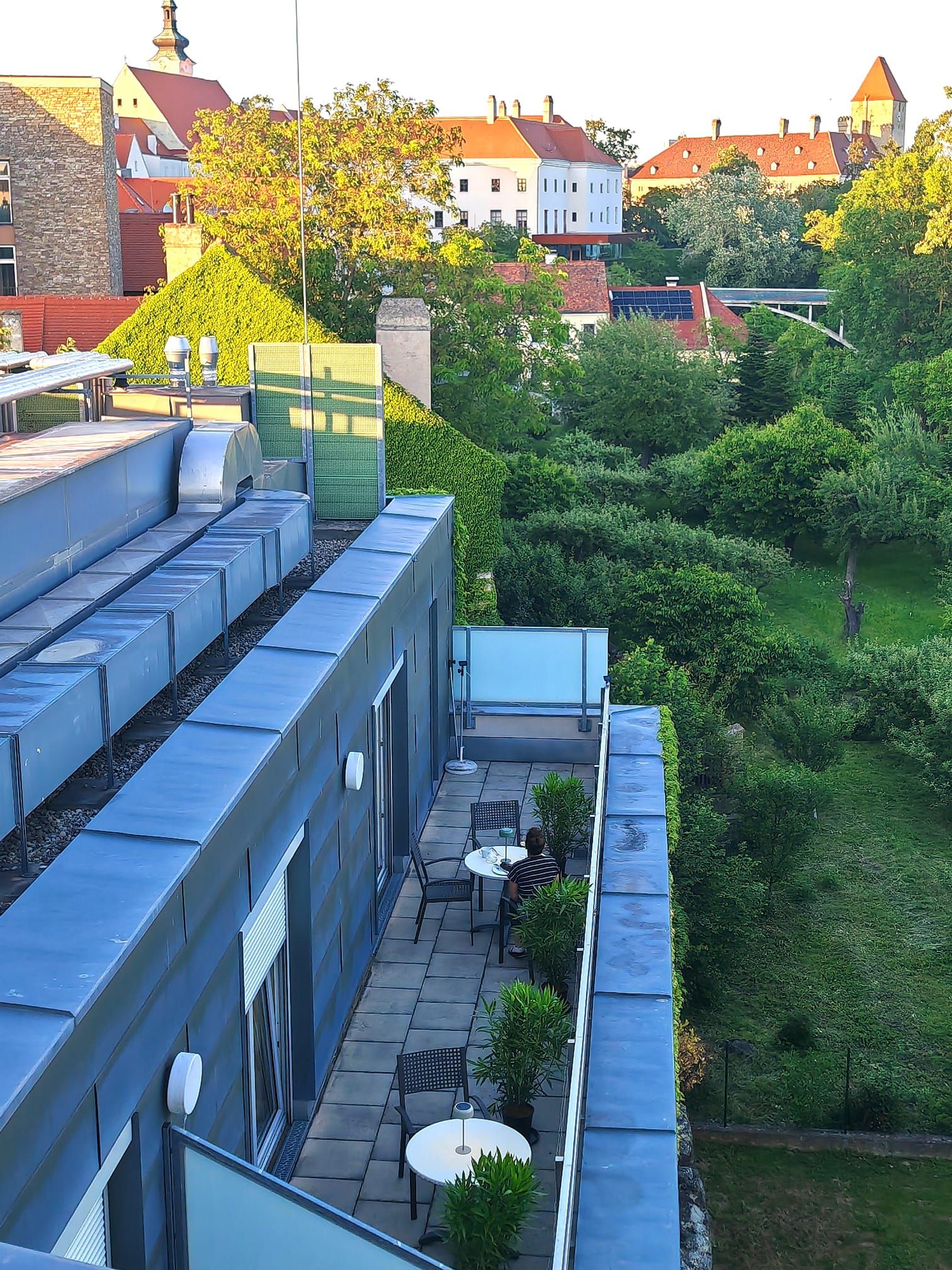 Blick von oben auf eine Dachterrasse mit Tischen und Stühlen, umgeben von grünen Pflanzen. Im Hintergrund sind Gebäude mit roten Dächern und Bäume zu sehen.