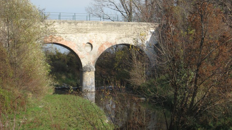Alte Steinbrücke über einen kleinen Fluss, umgeben von Bäumen im Herbst.