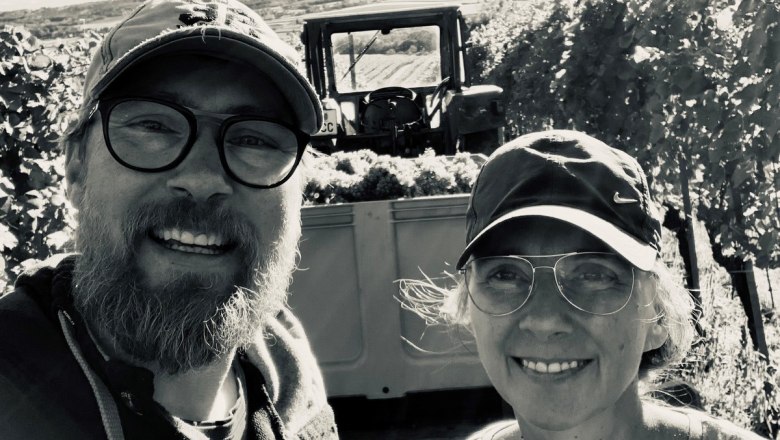 Ein Winzerpaar l&auml;chelt in die Kamera, im Hintergrund ein Traktor mit Weintrauben.