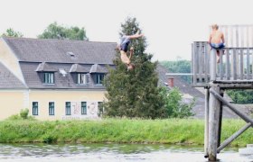 Ein Junge springt kopf&uuml;ber von einem Holzsteg in einen Teich, w&auml;hrend ein anderer Junge zuschaut. Im Hintergrund ist ein Geb&auml;ude mit der Aufschrift 'Gasthaus' zu sehen.