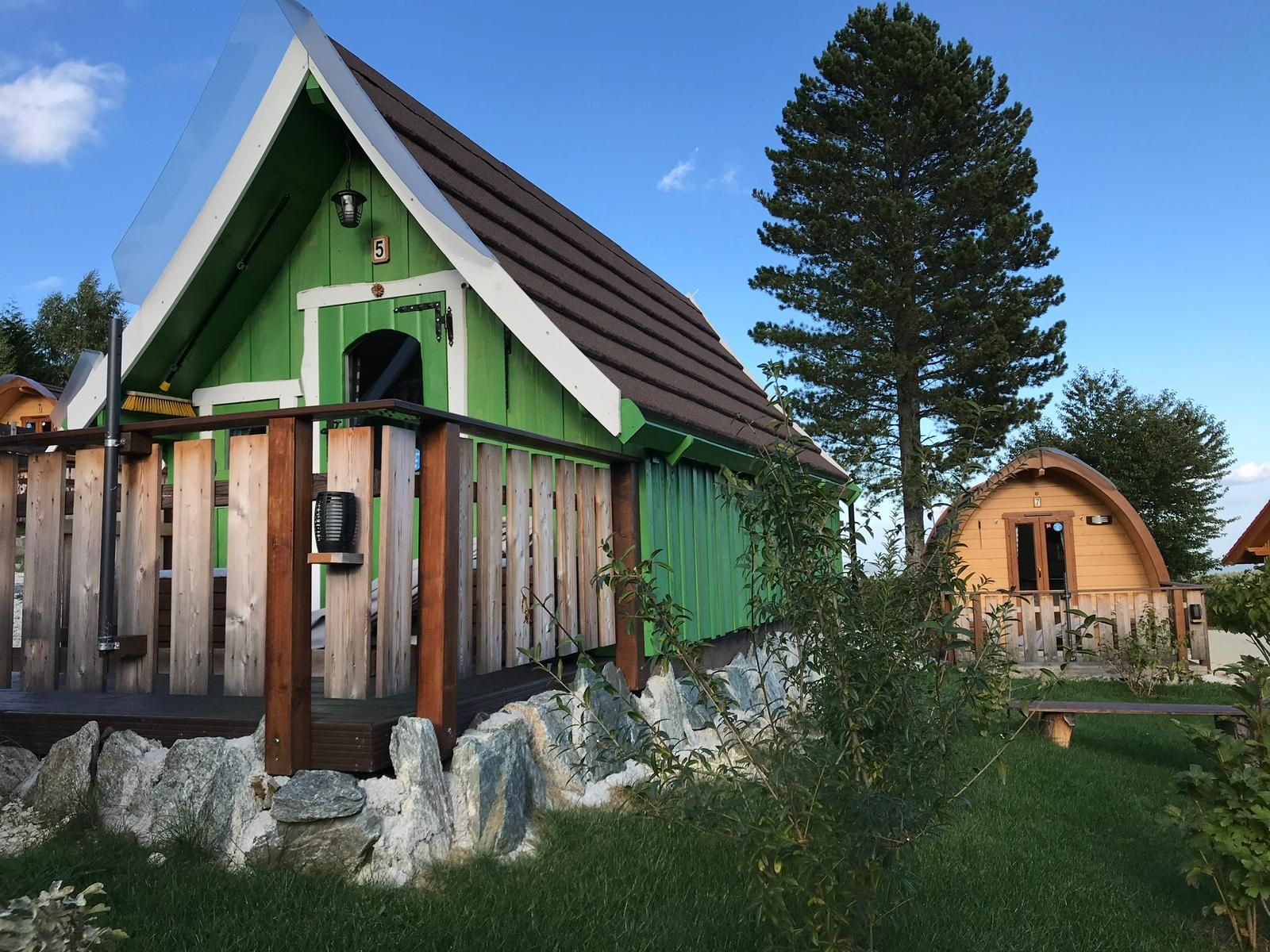 Zwei kleine Holzhütten im Glamping-Stil mit grüner und brauner Fassade, umgeben von Bäumen und Gras.