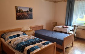 Ein einfach eingerichtetes Schlafzimmer mit zwei Einzelbetten, einem Tisch mit Stuhl und einem Fenster mit Vorh&auml;ngen.