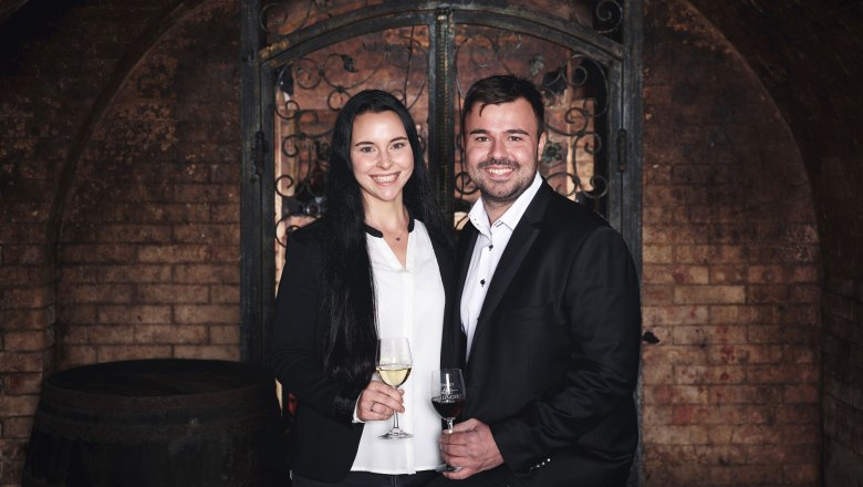 Ein Mann und eine Frau in eleganter Kleidung stehen in einem Weinkeller mit Weingläsern in der Hand.