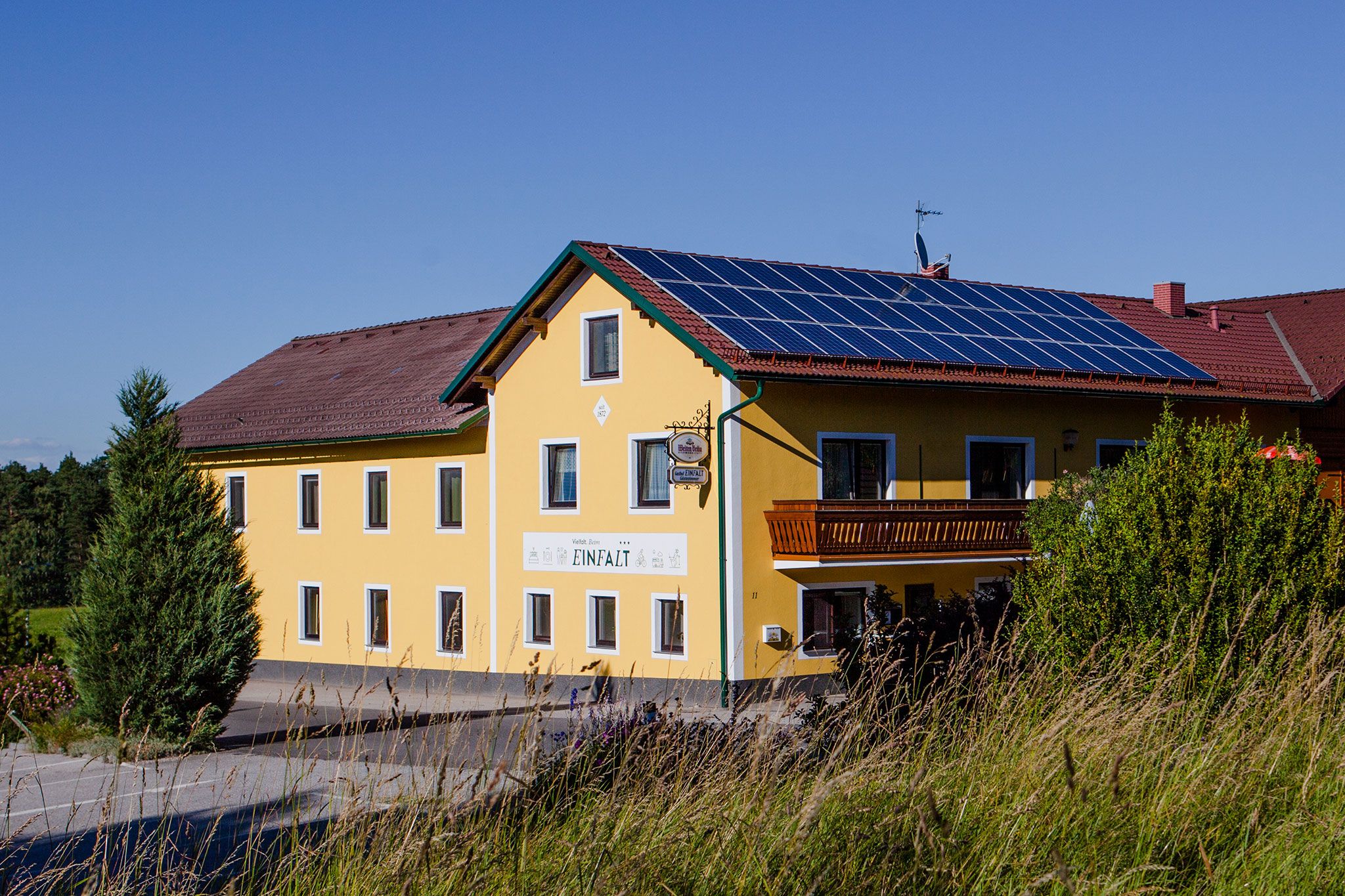 Ein gelbes Gasthaus mit Solarpanelen auf dem Dach und einem Balkon, umgeben von Bäumen und Gras.