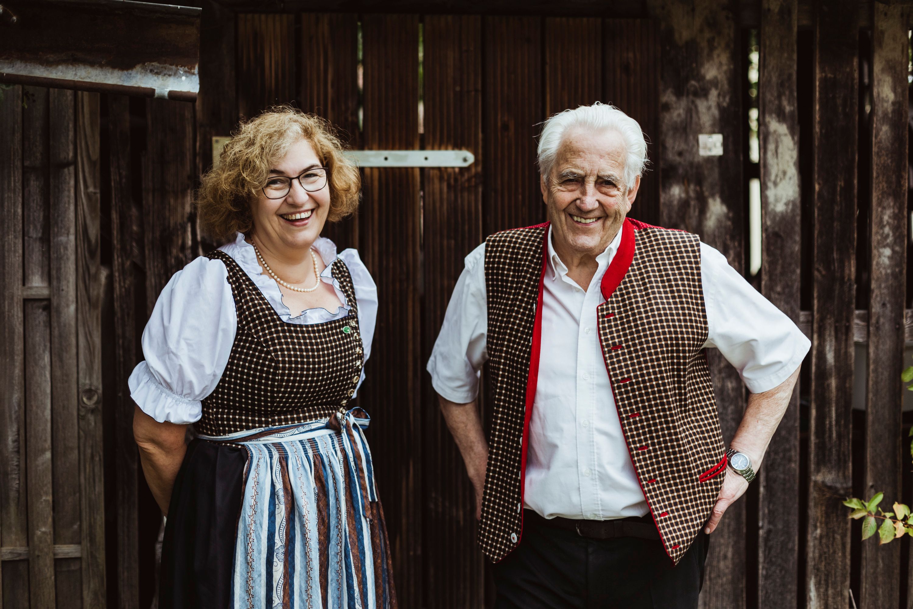 Zwei Personen in traditioneller Tracht vor einer Holzwand, lächelnd.