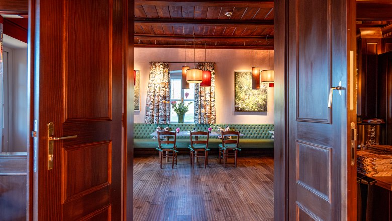 Blick durch geöffnete Holztüren in einen gemütlichen Raum mit Holzboden, Tisch und Stühlen, dekoriert mit Lampen und Vorhängen.