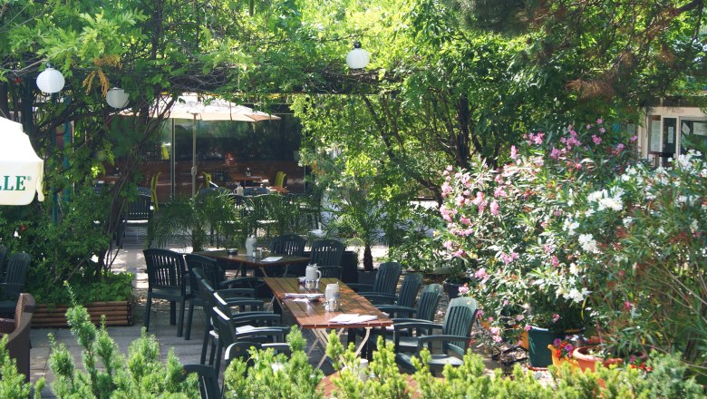 Ein grüner Garten mit Tischen und Stühlen, umgeben von Bäumen und blühenden Sträuchern.