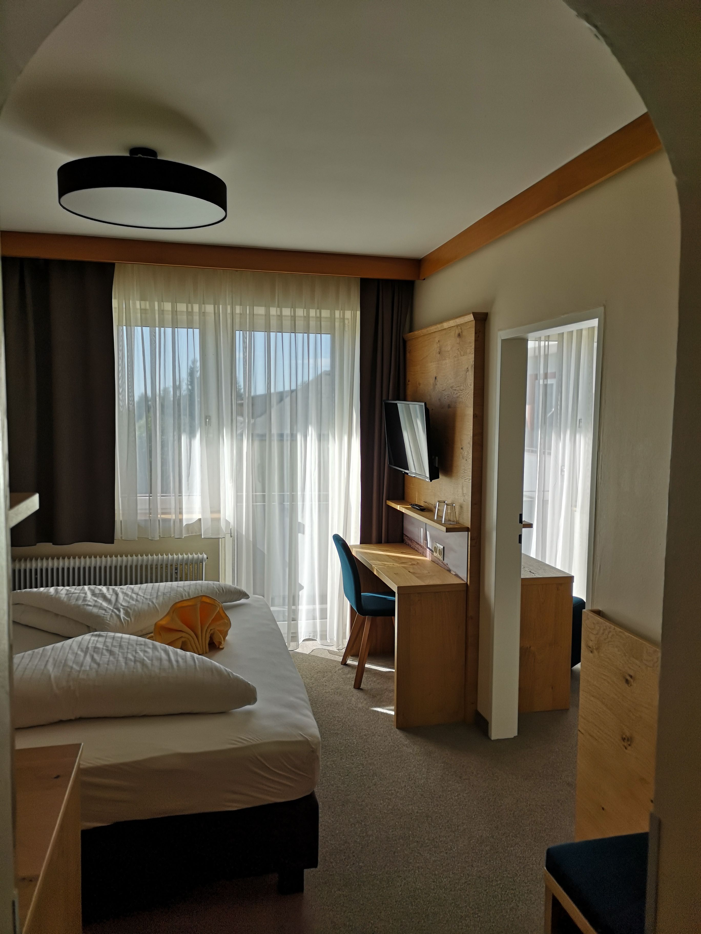 Ein modernes Doppelzimmer mit einem Bett, Schreibtisch, Stuhl und Fernseher. Große Fenster mit Vorhängen lassen Tageslicht herein.