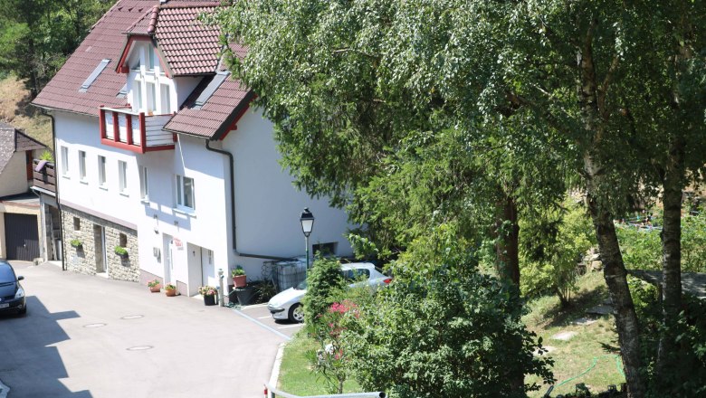 Au&szlig;enansicht eines Hauses mit rotem Dach und wei&szlig;en W&auml;nden, umgeben von B&auml;umen und einem Garten.