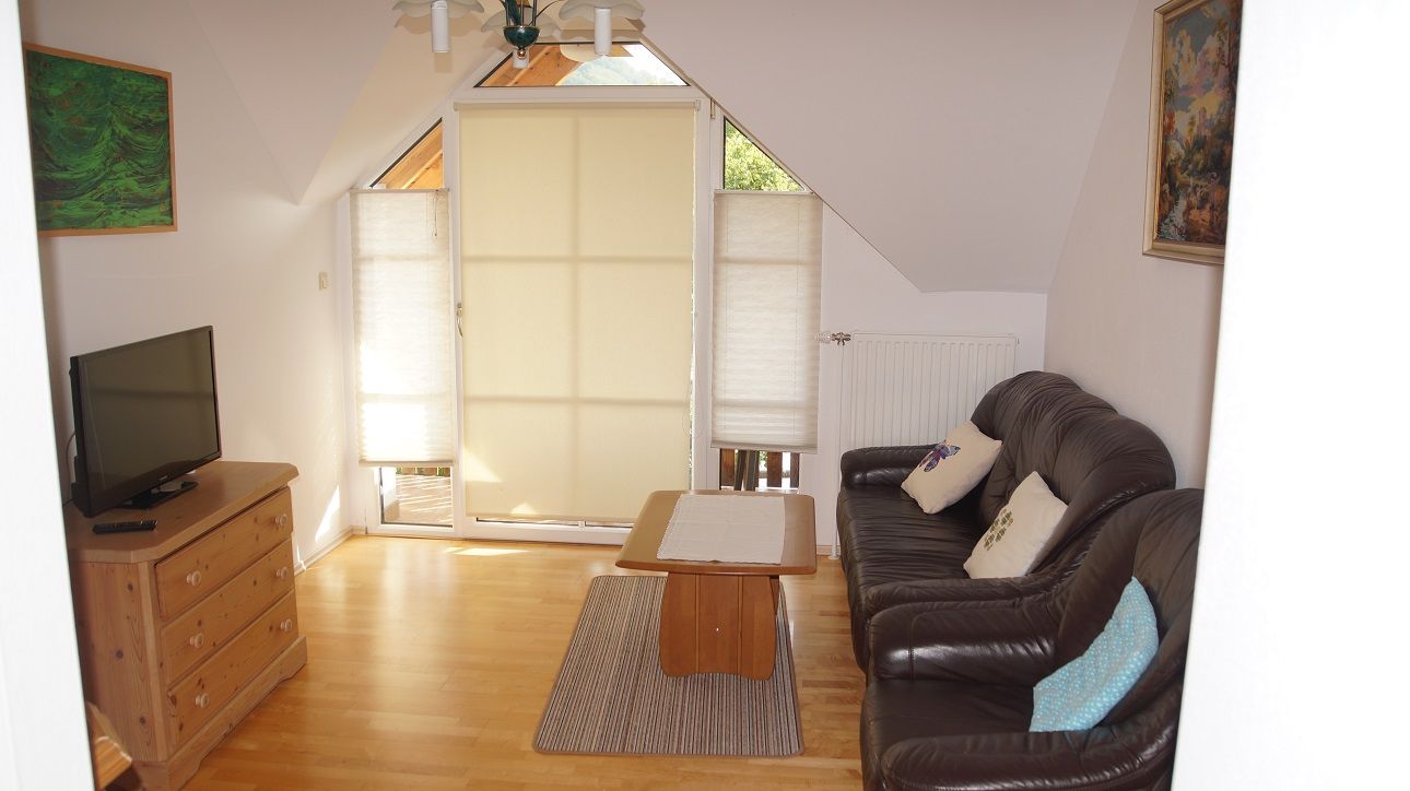 Wohnzimmer mit Ledersofa, Holztisch, Kommode und Fernseher. Fenster mit Jalousien, Bilder an den Wänden.