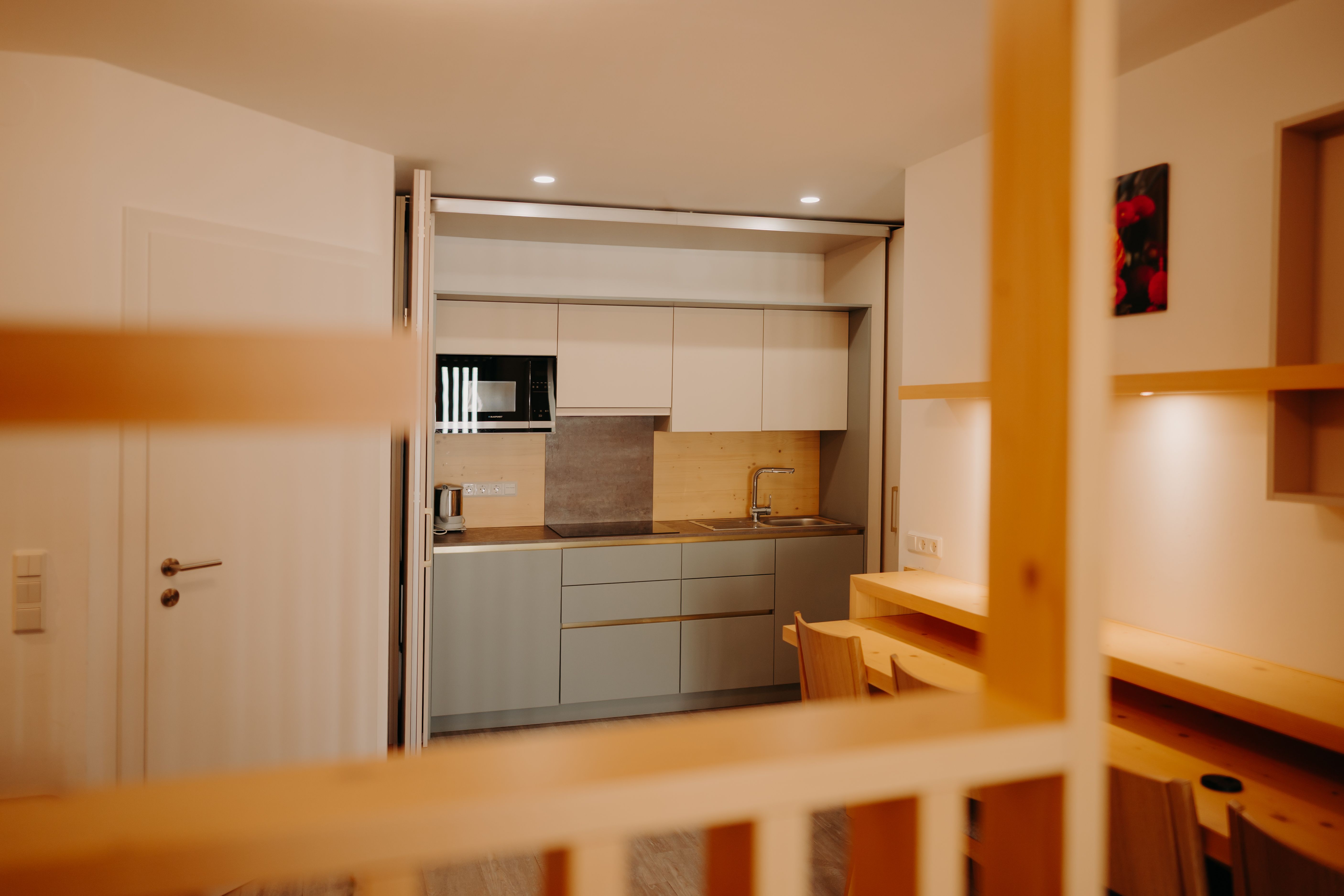 Moderne Küchenzeile in einem Apartment in hellen Farbtönen. Man sieht eine Spüle, eine Mikrowelle, eine große Herdplatte und einen Wasserkocher. Die Küche kann hinter einer verschiebbaren Trennwand versteckt werden. Vor der Küche sieht man einen Tisch an der Wand und 4 Sessel.