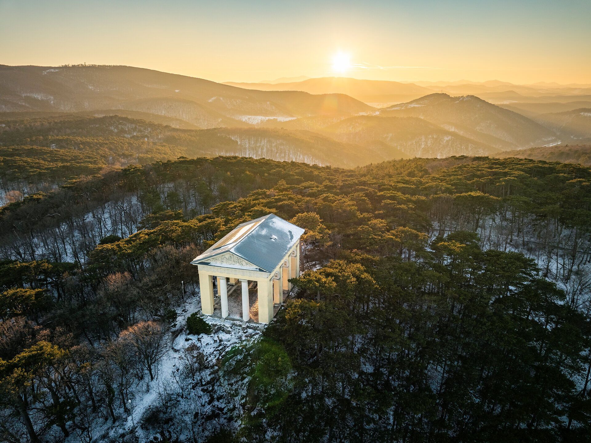 Drohnenaufnahme des Husarentempels im winterlichen Wienerwald, mit verschneiten Baumkronen und Blick über die hügelige Landschaft im warmen Licht des Sonnenaufgangs.
