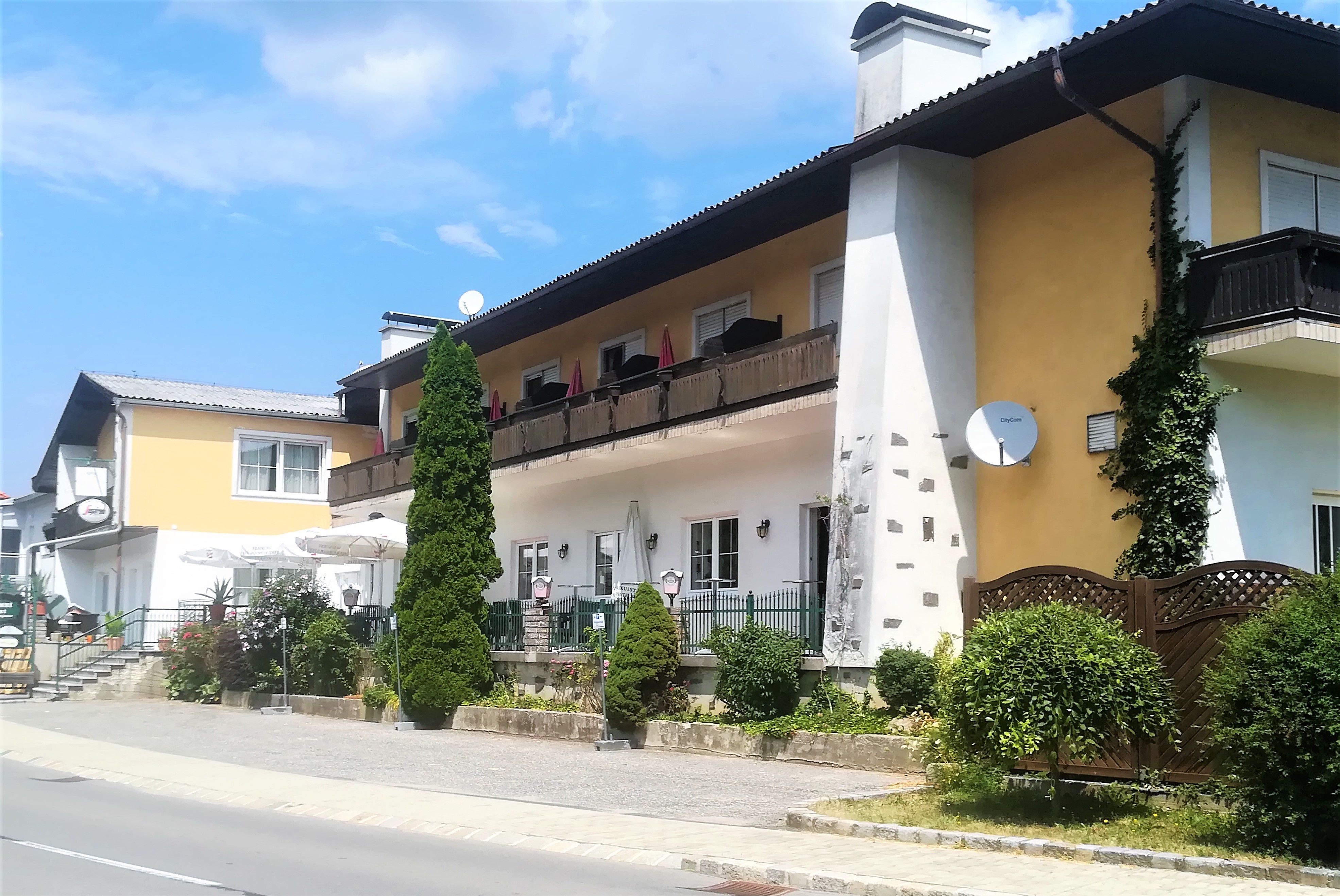 Ein zweistöckiges Gebäude mit Balkon und Terrasse, umgeben von Pflanzen und Bäumen, bei sonnigem Wetter.