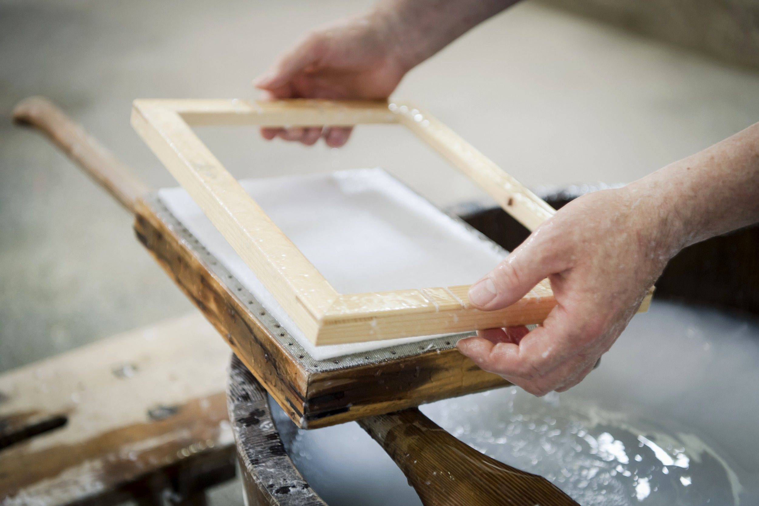 Nahaufnahme von Händen, die einen Holzrahmen über einer Papiermasse in einer traditionellen Papiermühle halten.