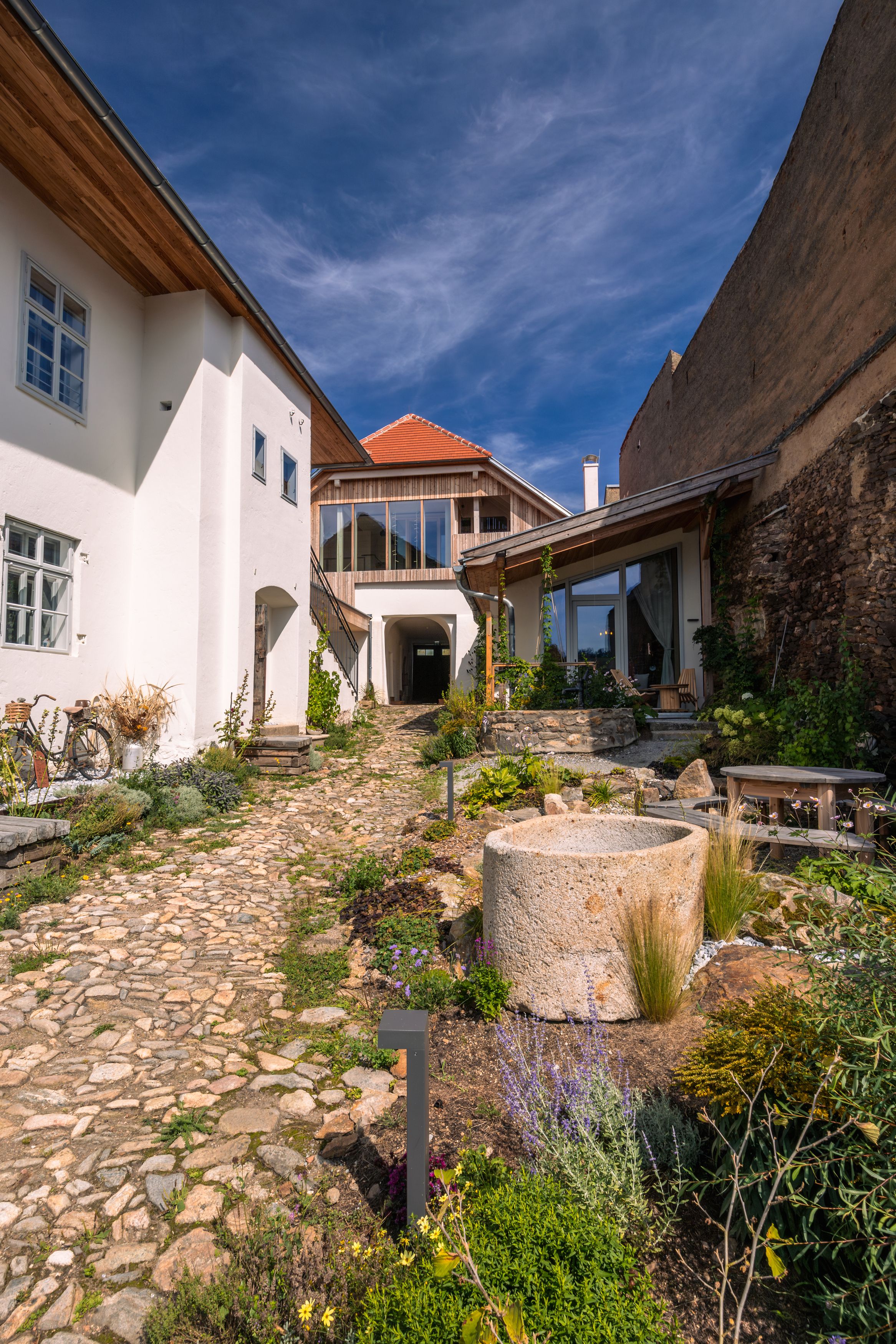 Innenhof mit gepflastertem Weg, Pflanzen und modernen Gebäuden unter blauem Himmel.