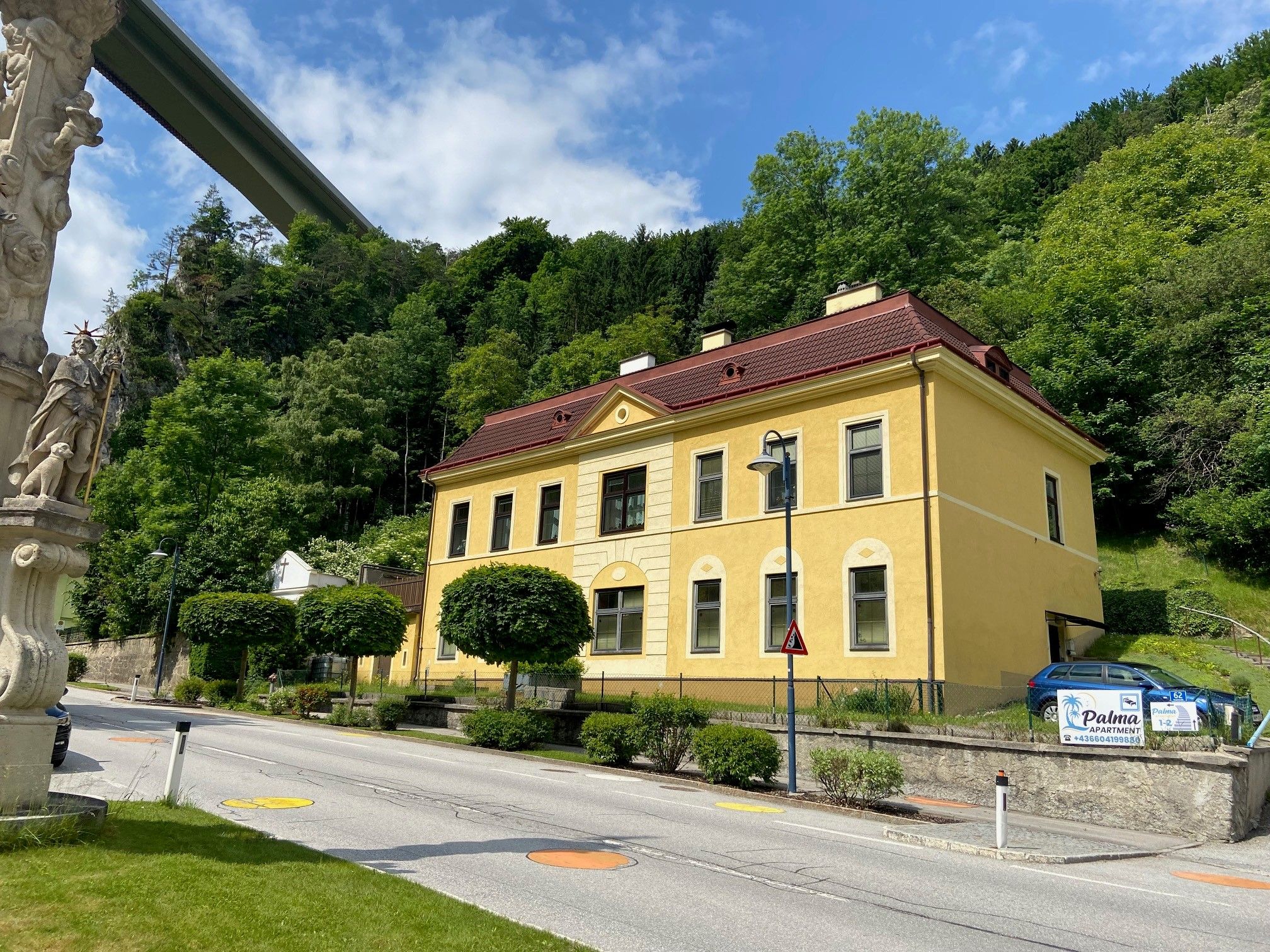 Gelbes Gebäude mit rotem Dach neben einer Straße, umgeben von Bäumen und einer Statue im Vordergrund.