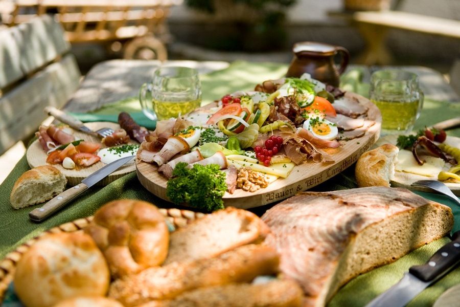 Ein reich gedeckter Tisch mit einer Auswahl an Wurst, Käse, Brot und Bierkrügen im Freien.