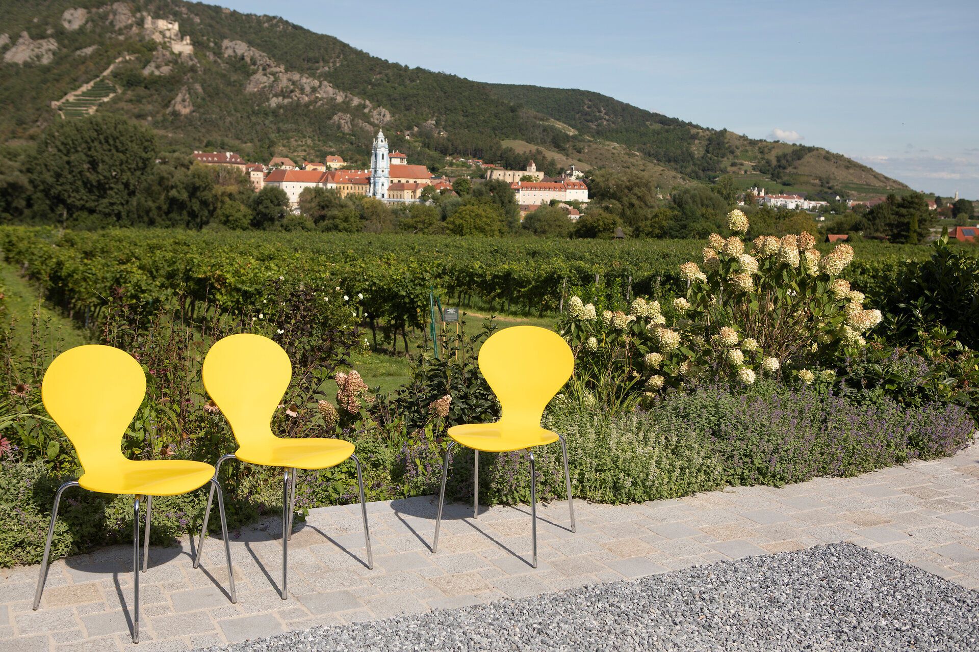 Die strahlend gelben Stühle laden dazu ein, die atemberaubende Aussicht auf die sanften Hügel und Weinberge zu genießen. Umgeben von üppigem Grün und blühenden Pflanzen, vermittelt dieser Ort ein Gefühl von Ruhe und Entspannung. Hier, wo die Natur und die Kultur harmonisch verschmelzen, wird jeder Moment zum unvergesslichen Erlebnis.
