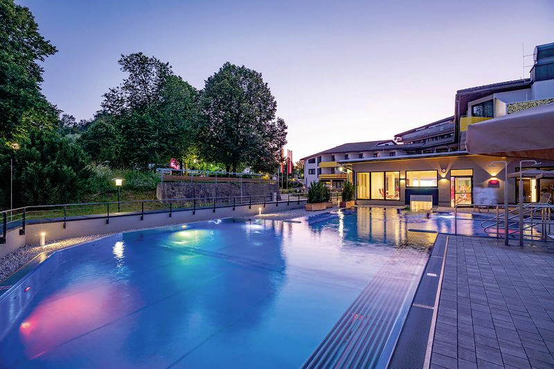 Beleuchtete Poolanlage bei Nacht mit Gebäuden im Hintergrund.