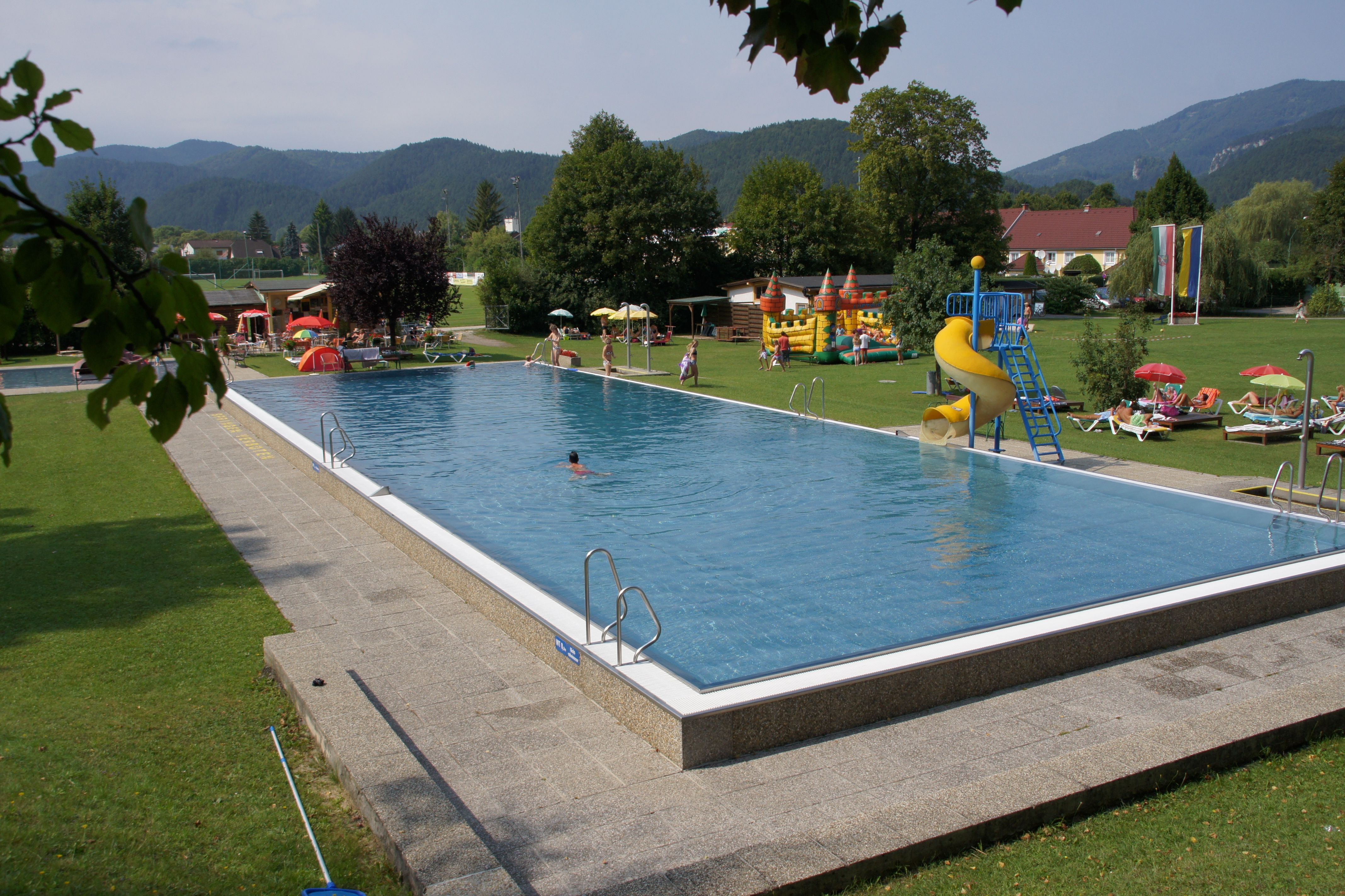 Ein Freibad mit Schwimmbecken, Rutsche und Liegewiese, umgeben von Bäumen und Bergen.