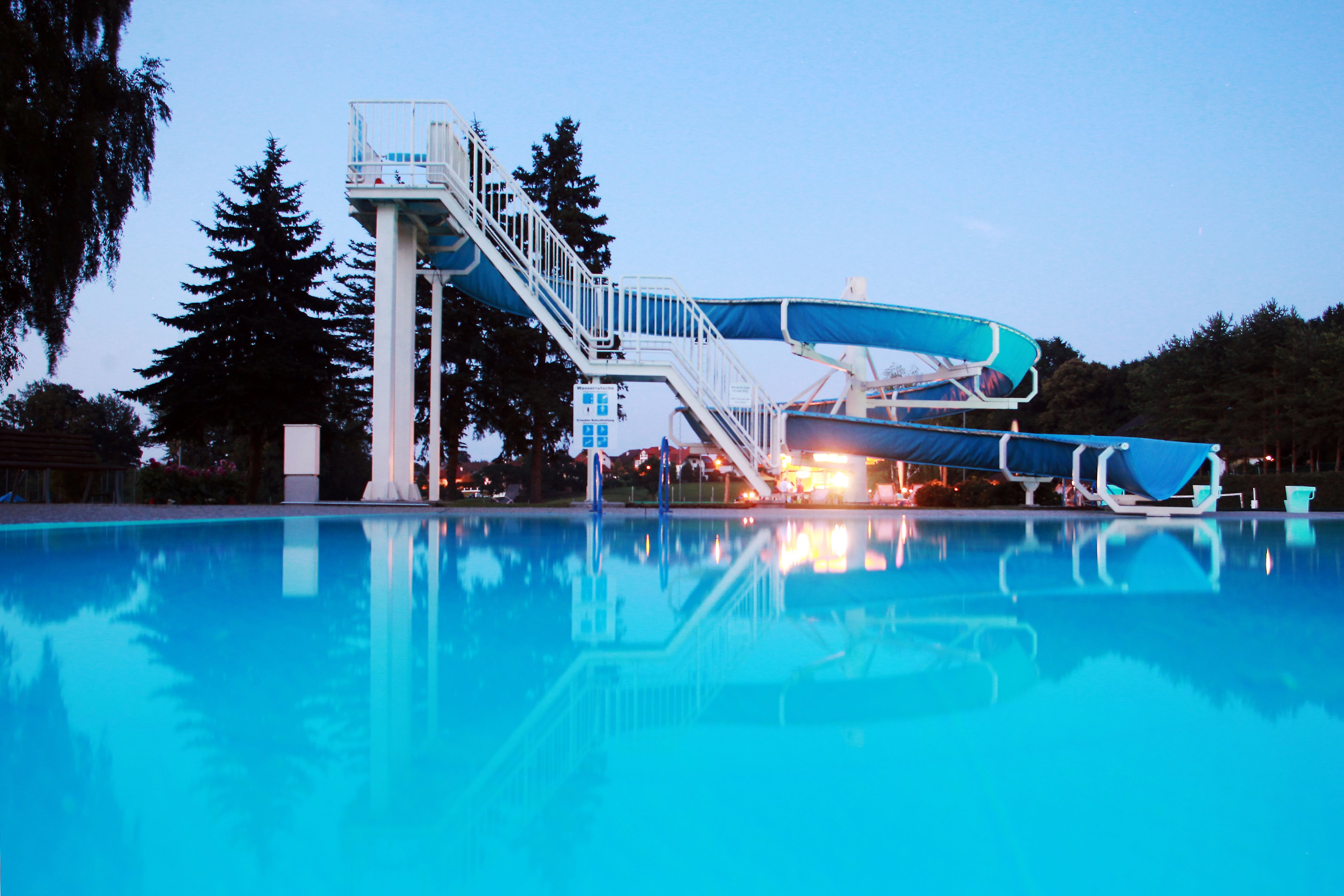 Ein Freibad mit einer großen blauen Wasserrutsche und einem klaren Pool im Vordergrund, umgeben von Bäumen bei Dämmerung.