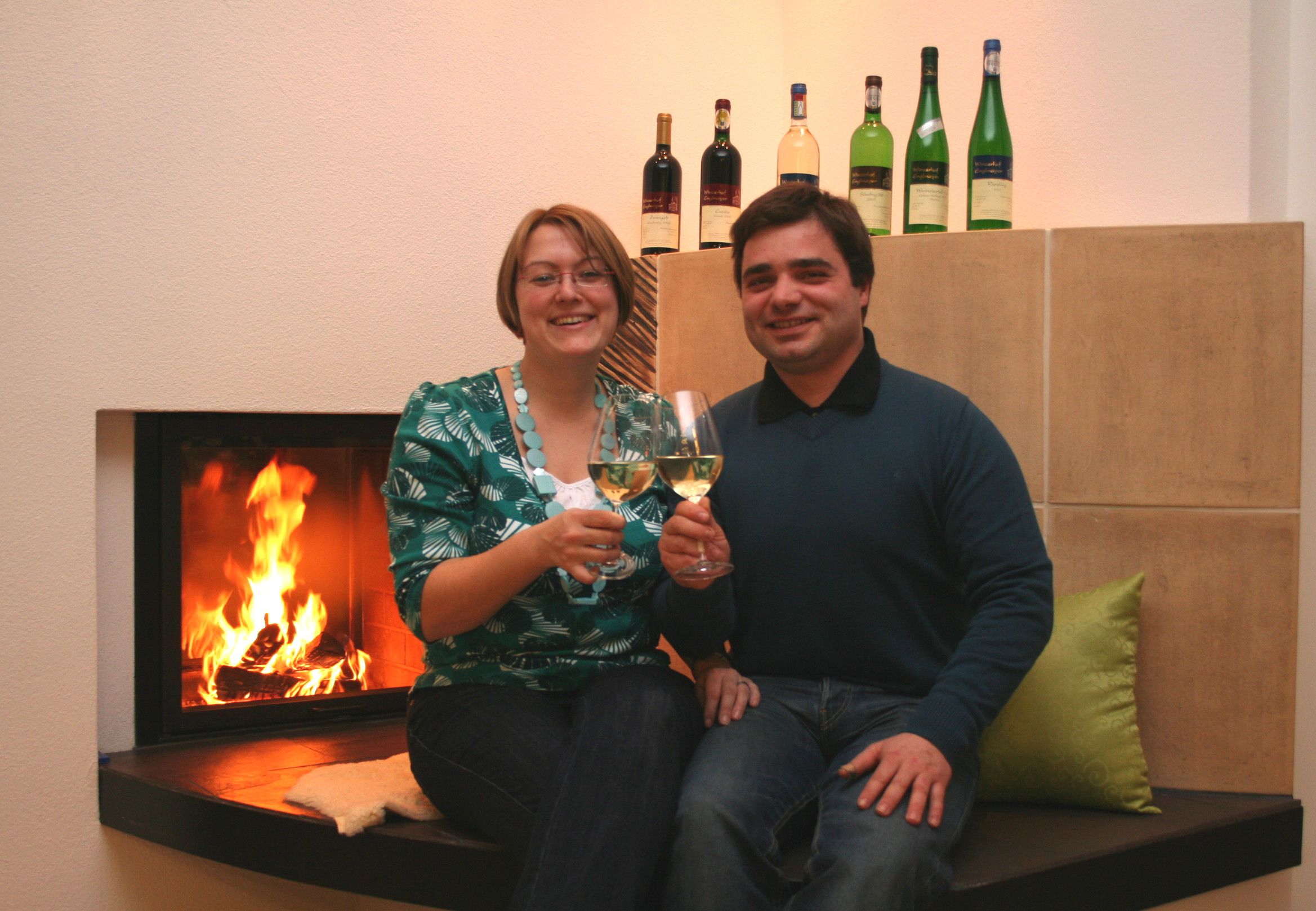 Ein Paar sitzt vor einem Kamin und stößt mit Weingläsern an.