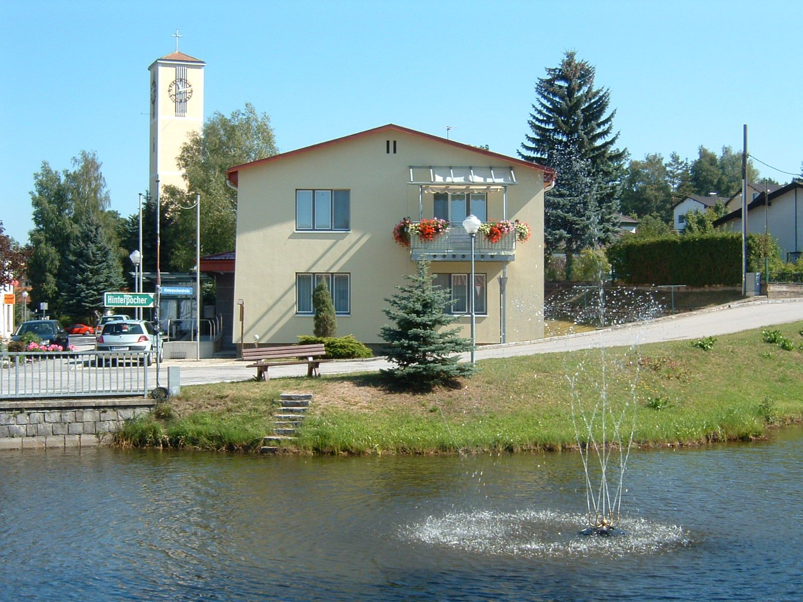 Ein gelbes Gebäude mit Blumenkästen und einem Glockenturm im Hintergrund, davor ein Teich mit Springbrunnen.