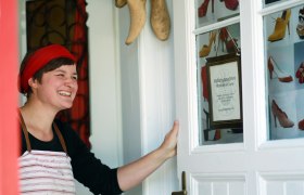 Eine Frau mit rotem Stirnband und Schürze steht lächelnd an einer Tür mit Schuhbildern und einem Schild der Maßschuhmacherei Pfaffenlehner Doris.