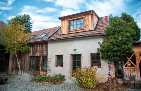 Rustikales Haus mit Ziegeldach und Holzverkleidung, umgeben von Pflanzen und Kopfsteinpflaster.