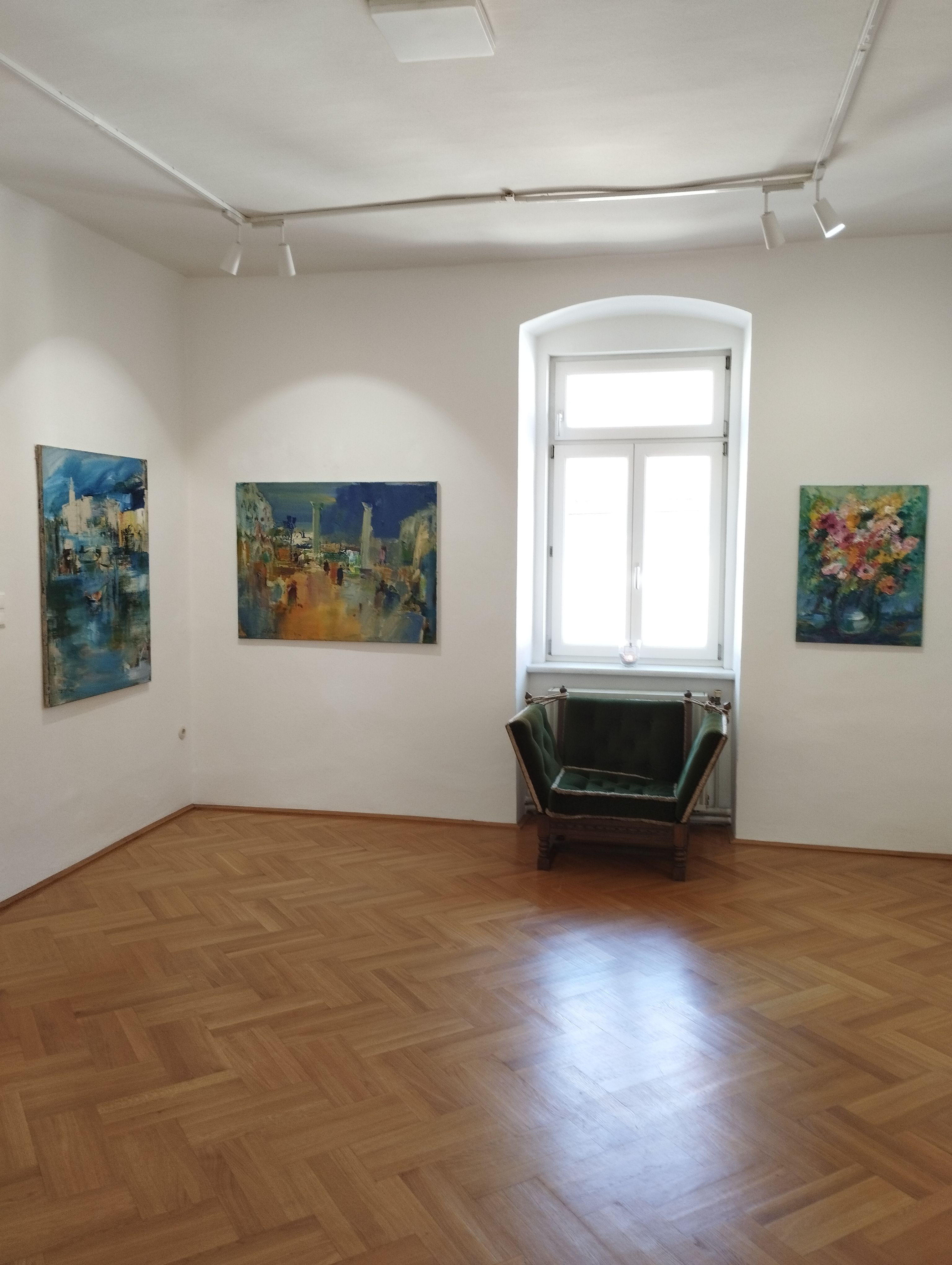 Innenansicht einer Galerie mit drei Gemälden an den Wänden und einem grünen Sessel vor einem Fenster.