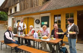 Gruppe von Menschen sitzt an einem Tisch vor einem rustikalen Geb&auml;ude und genie&szlig;t eine Mahlzeit im Freien.