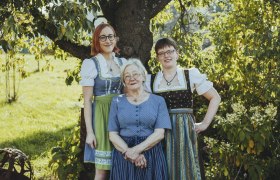 Drei Frauen in traditioneller Tracht stehen lächelnd unter einem Baum im Freien.