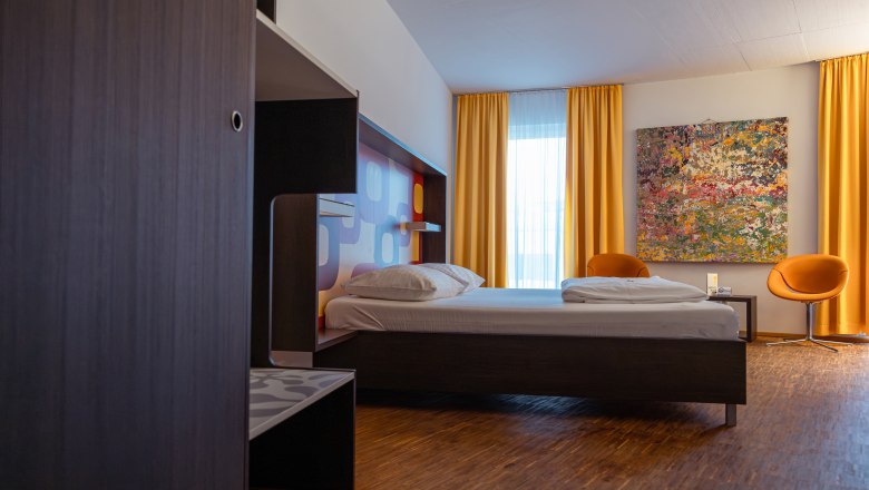 Modernes Hotelzimmer mit Bett, gelben Vorh&auml;ngen und buntem Gem&auml;lde an der Wand.