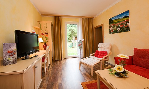 Gem&uuml;tliches Zimmer mit roten Akzenten, Fernseher, Sofa, Sessel und Blick auf den Garten durch eine Glast&uuml;r.