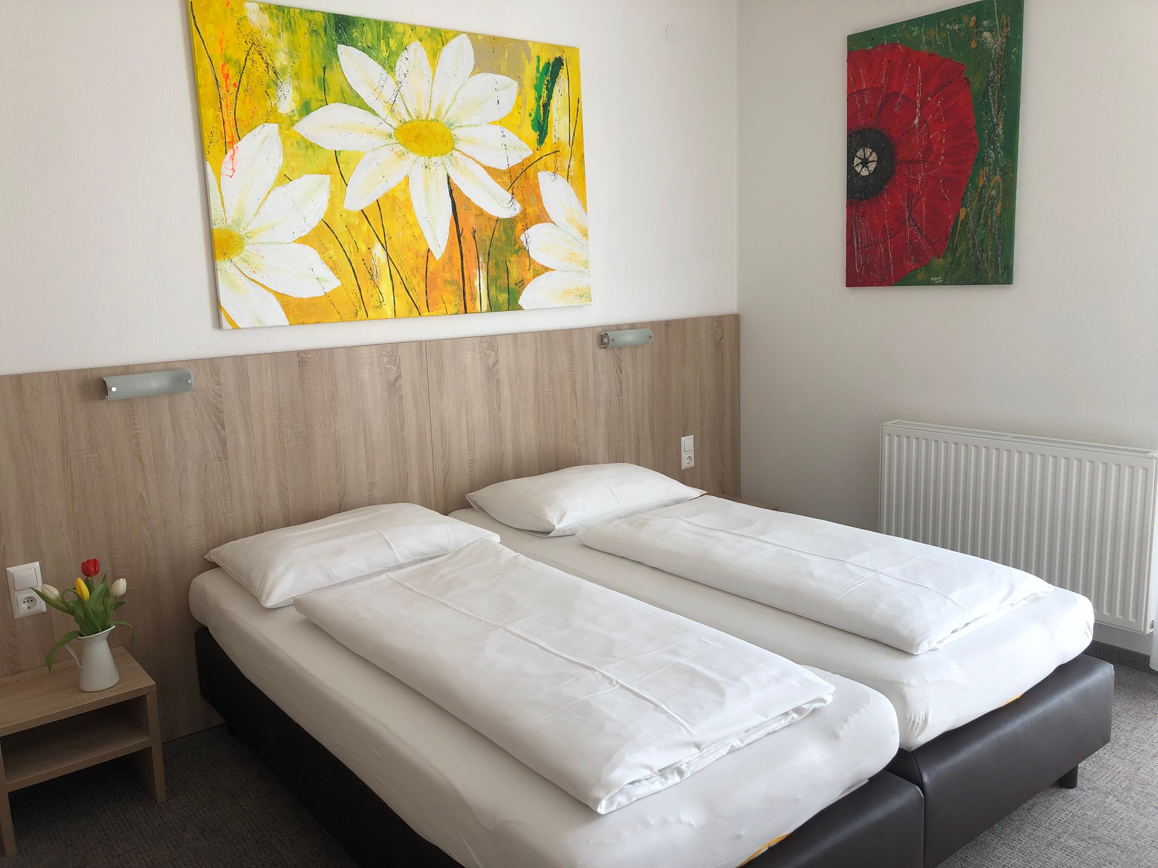 Ein schlichtes Hotelzimmer mit zwei Einzelbetten, einem Nachttisch mit Blumen und zwei bunten Gemälden an der Wand.