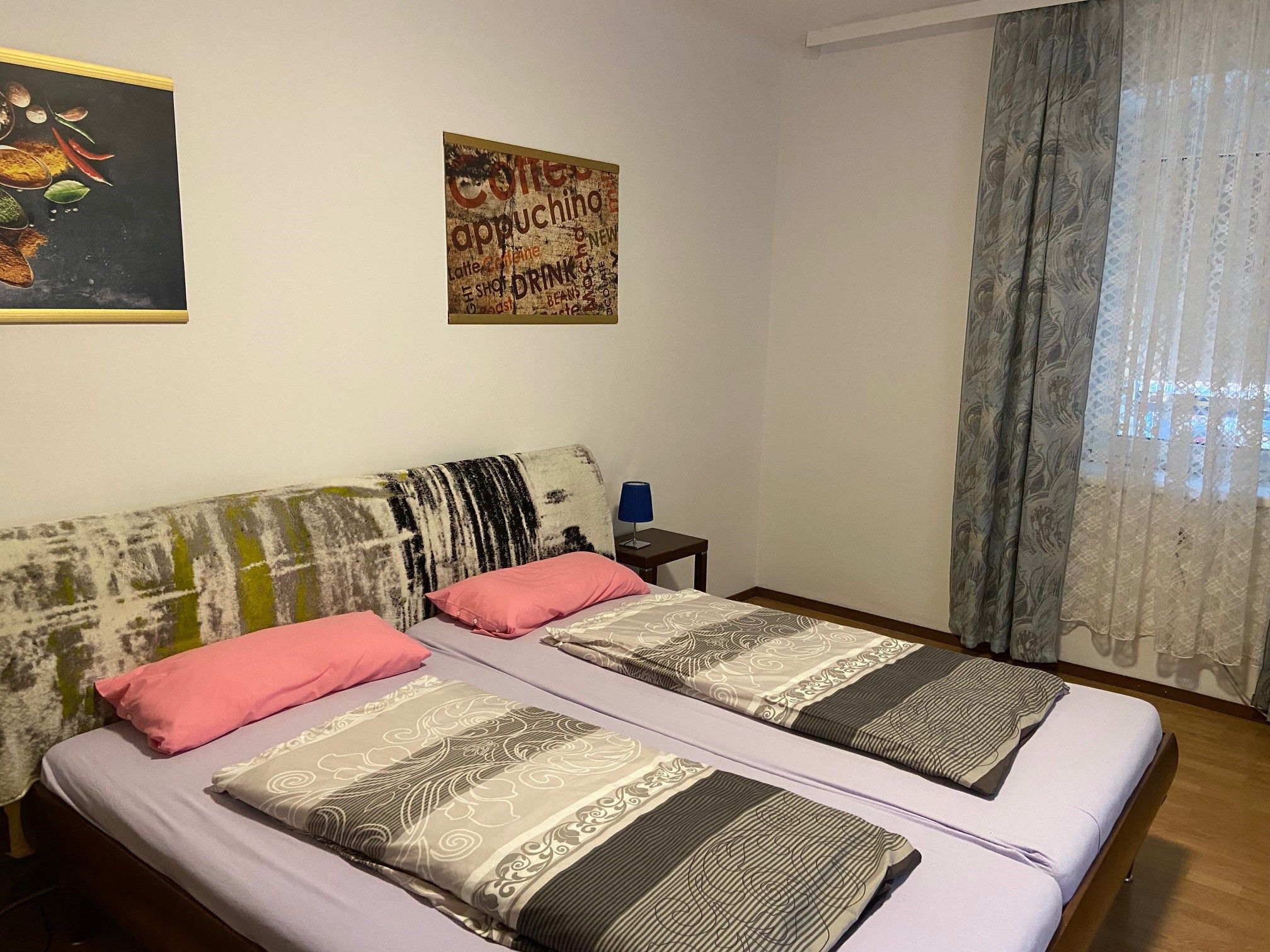 Schlafzimmer mit Doppelbett, zwei Kissen, zwei Decken, Wandbilder und Fenster mit Vorhängen.