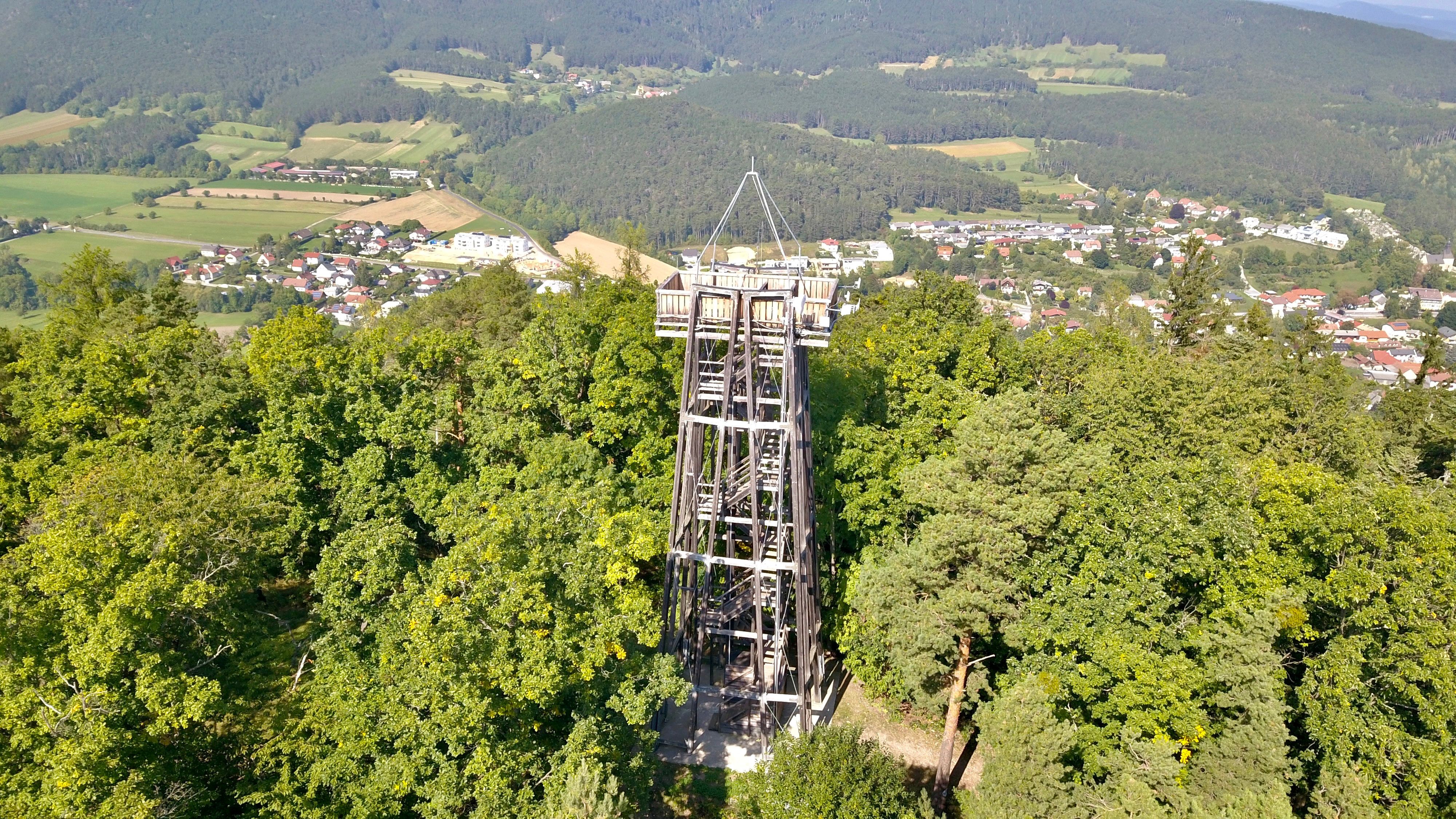 Aussichtsturm inmitten von Bäumen mit Blick auf ein Dorf und bewaldete Hügel im Hintergrund.