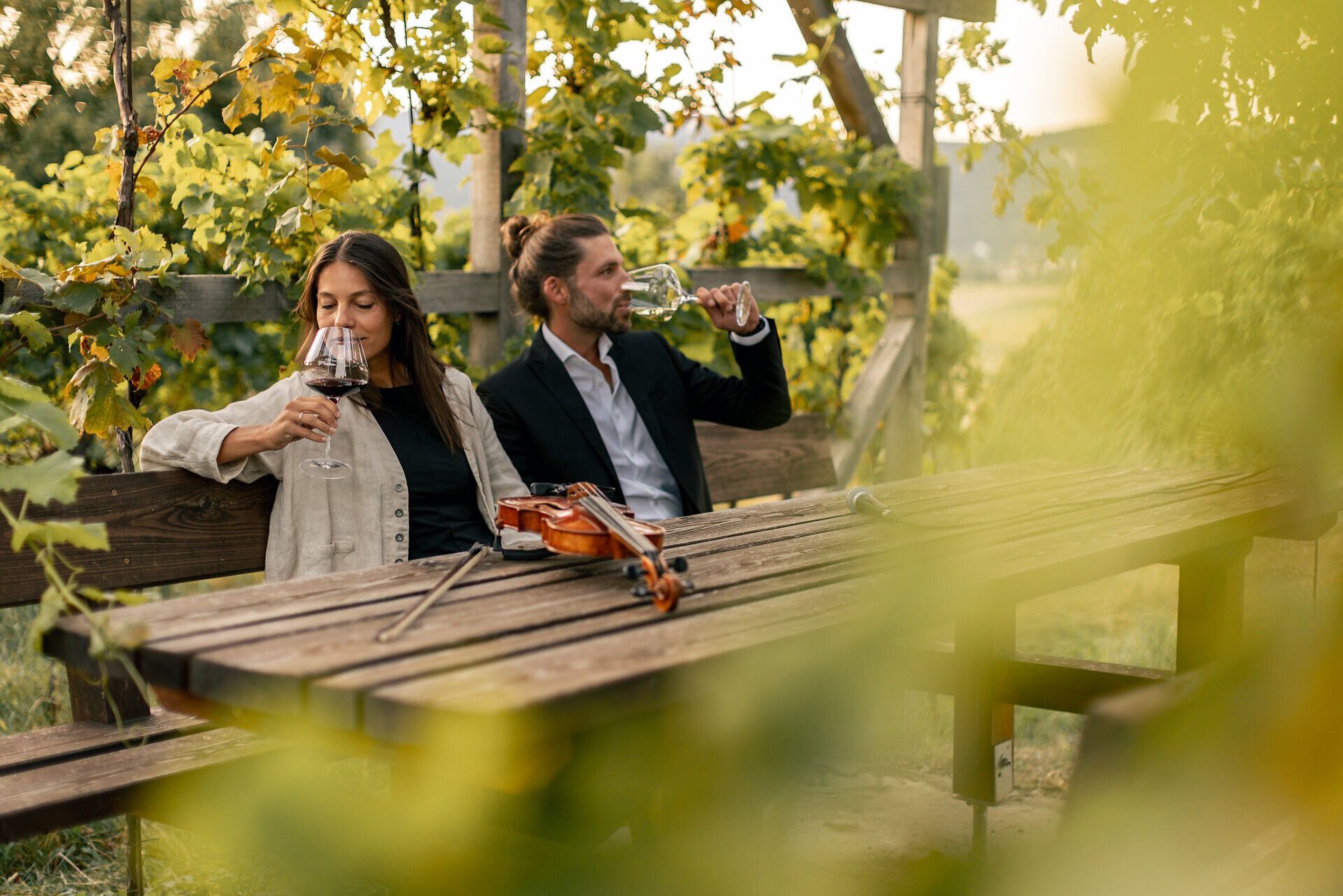 Zwei Personen sitzen an einem Holztisch im Weinberg und verkosten Wein; eine Geige liegt auf dem Tisch, Reben im Vordergrund.