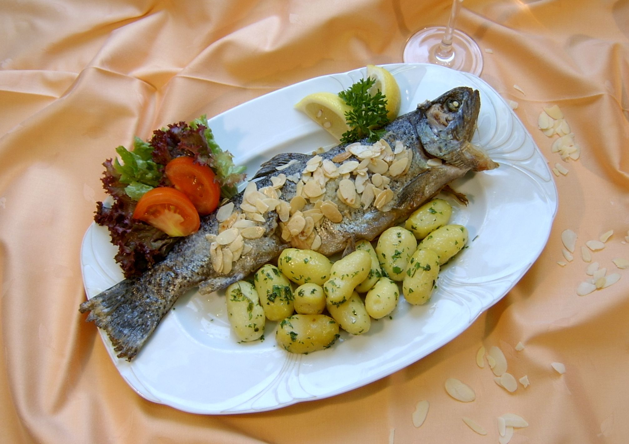 Ein Teller mit einer gebratenen Forelle, bedeckt mit Mandeln, serviert mit Kartoffeln, Salat und Zitronenscheiben auf einem orangefarbenen Tuch.