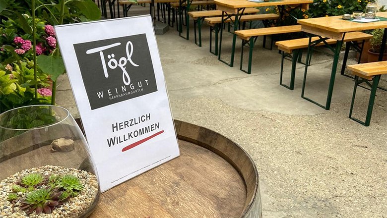 Ein Schild mit der Aufschrift 'Weingut T&ouml;gl' und 'Herzlich Willkommen' steht auf einem Weinfass. Im Hintergrund sind Biertische und Pflanzen zu sehen.