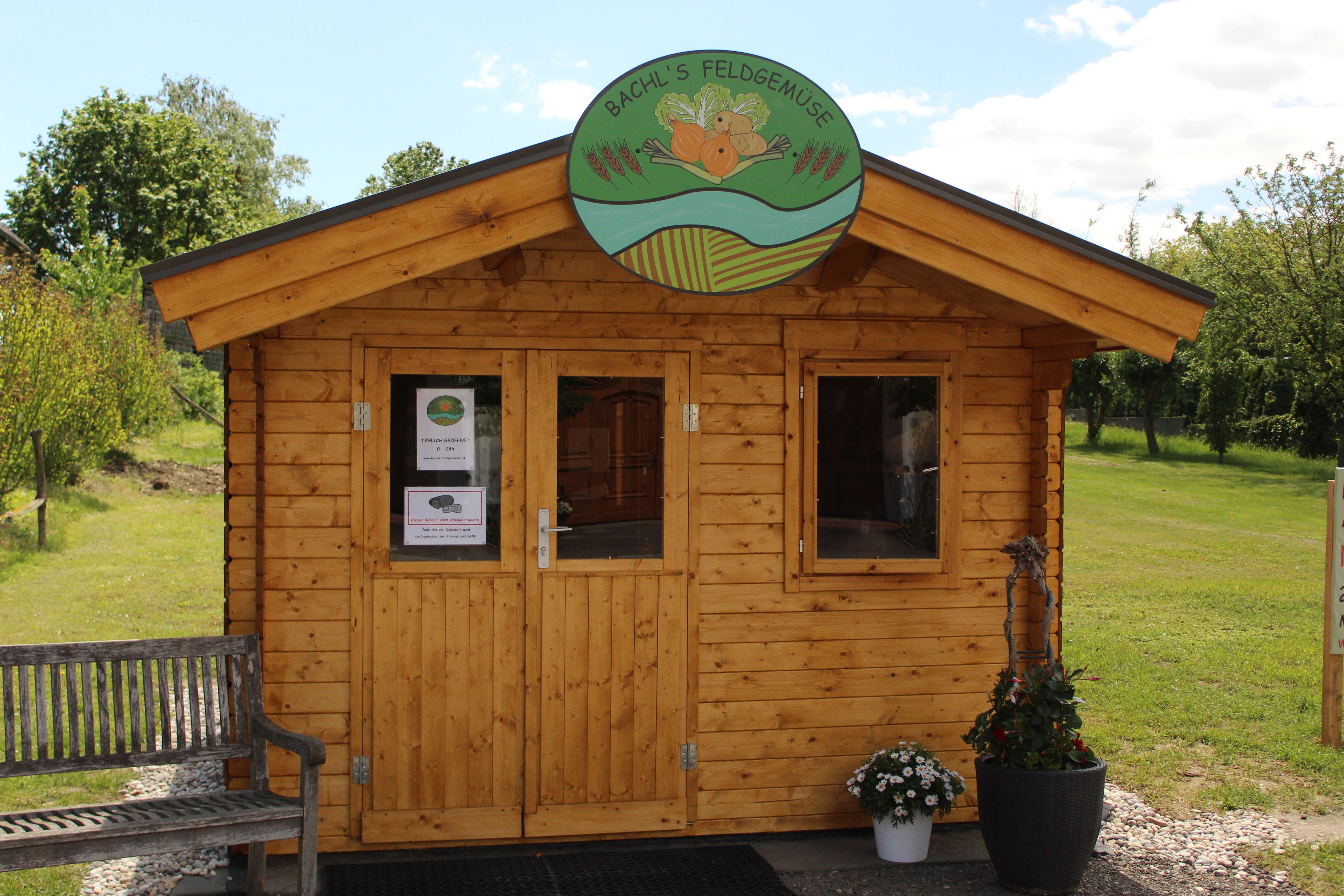 Holzhütte mit Schild 'Bachl's Feldgemüse' und Blumen davor.