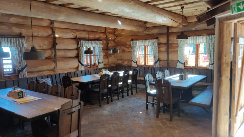 Innenansicht einer rustikalen Holzh&uuml;tte mit Holztischen und St&uuml;hlen, dekoriert mit Vorh&auml;ngen an den Fenstern.