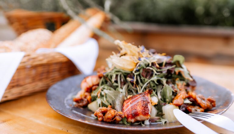 Ein Teller mit Salat und gegrilltem Fleisch auf einem Holztisch, im Hintergrund ein Brotkorb.
